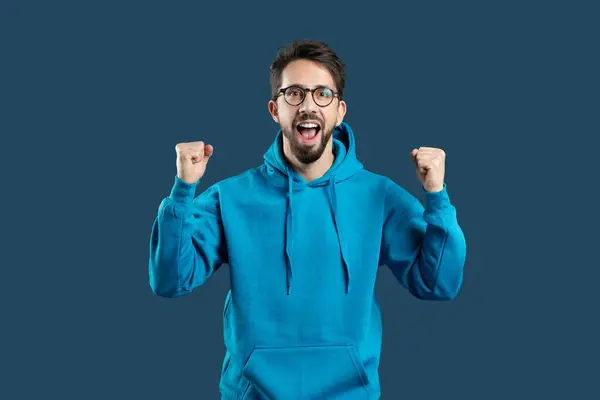 stock image A man in a blue hoodie with his fists raised celebrating success, showing positive emotions isolated on blue studio background