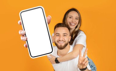 Neşeli genç bir çift yan yana duruyor, ikisi de kameraya doğru cep telefonu tutuyorlar. Telefon ekranına bakıyorlar, muhtemelen bir selfie ya da video görüşmesi yapıyorlar..