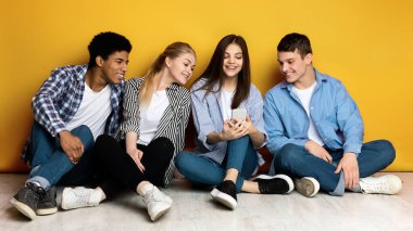 Dört arkadaş, çok ırklı gençler, parlak sarı bir duvara yaslanarak yerde yan yana oturuyorlar. Bir tanesinin elinde akıllı telefon var ve hepsi ekrana bakıyor.