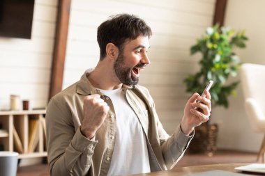 Sağ elinde cep telefonu tutan bir adam görülüyor, parmaklarını cihaza dolamış. Eli göğüs hizasında ve ekrana odaklanmış gibi görünüyor..