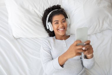 İspanyol kadın elinde telefonuyla yatakta uzanıyor, kulaklık takıyor ve müzik dinliyor. Rahatlamış görünüyor ve telefonuyla çalan müziğe odaklanmış durumda..