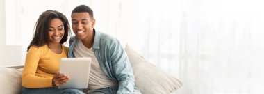 Kanepede tablet kullanan Afro-Amerikan çift web-afiş alanı ile samimi bir görüntü sunuyorlar.