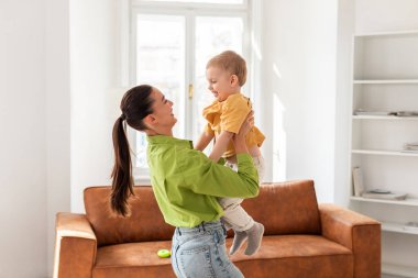 Bir kadın, her ikisi de gülümseyen bir bebeği neşeyle havaya kaldırır. Çocuk kolları açık, evdeki anne ile eğlenceli etkileşimin keyfini çıkarıyorlar.