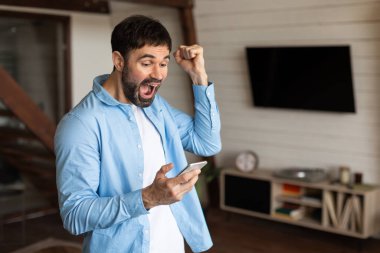 Mavi gömlekli bir adam elinde bir cep telefonu tutarken ayakta duruyor, el kol hareketi yapıyor, neşeyi paylaşıyor, başarıyı kutluyor, ev içini.