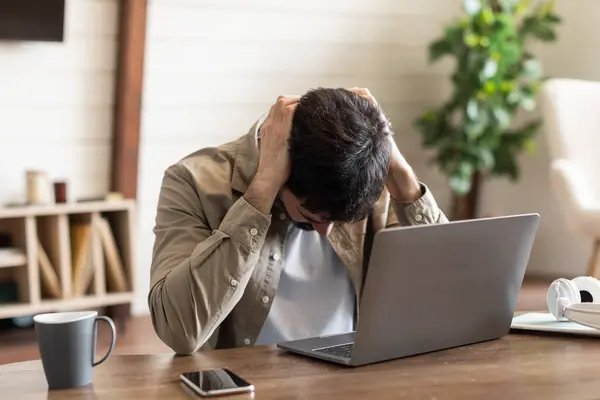 stock image A young man sits at a wooden desk, his hands clutching his head in a gesture of frustration or overwhelm. An open laptop, a mobile phone, and a cup of coffee accompany him