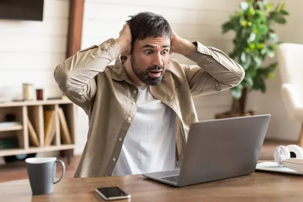 stock image A man is seated at a wood table, hands on his head with an expression of shock and disbelief as he looks at his laptop screen.