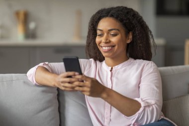 Mutlu genç siyah kadın akıllı telefon kullanıyor, koltukta tek başına oturuyor, mesajlaşıyor ya da web sörfü yapıyor, güzel mobil uygulama kullanıyor.