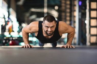 Bir adam spor salonunda şınav çekiyor. Antrenman kıyafeti giyiyor ve egzersiz rutinine odaklanmış durumda. Antrenmanında güç ve kararlılık gösteriyor..