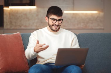 Bir adam kanepede oturuyor, dizüstü bilgisayarına dalmış. Ekrana dikkatle bakarken klavyeye basıyor, evden video çağrısı alıyor.