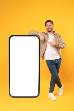 Açık renk ceket, kot pantolon ve beyaz spor ayakkabı giymiş neşeli bir adam beyaz ekranlı, büyük ve boş bir akıllı telefona yaslanıyor.