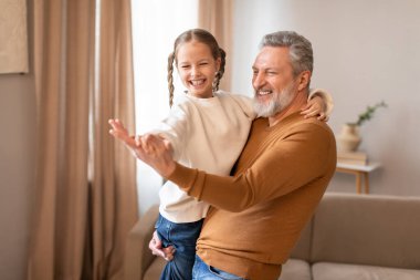 Senior man is standing, holding a little girl in his arms, grandfather dancing with his cute little granddaughter at home clipart