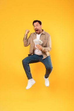 Bir adam elinde sıkıca para tutarken enerjik bir şekilde havaya zıplar. Coşkuyla zıplarken ayakları yerden kesiliyor..