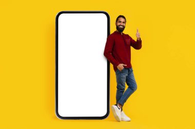 Kırmızı kazak ve mavi kot giyen neşeli Hintli bir adam büyük, boş bir akıllı telefon ekranının yanında duruyor ve sağ eliyle baş parmağını kaldırıyor.