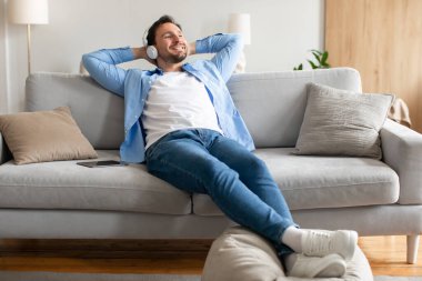 Bir adam rahat bir şekilde kanepede uzanıyor, kulaklıkları takılı, müzik dinliyor. Rahatlamış ve müzik çalmaya odaklanmış görünüyor. Oda rahat ve iyi aydınlatılmış, rahat bir atmosfer yaratıyor..