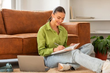 Bir kadın yerde oturuyor, dizüstü bilgisayar ve not defteri kullanıyor. O, görevleri üzerinde çalışırken odaklanmış görünüyor. Dizüstü bilgisayar odada hafif bir parıltı yayıyor..