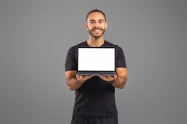 Bir adam ayakta duruyor, elinde boş beyaz ekranlı bir laptop var. Adamın eli dizüstü bilgisayarı sıkıca kavrarken, onun bakışı ekrana sabitlenir. Arkaplan basit ve göze çarpmayan.