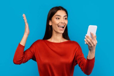 Kırmızı bluzlu bir kadın elinde bir cep telefonu tutuyor, ekranla meşgul gibi görünüyor. Cihazla etkileşime girerken duruşuna dikkat ediyor..