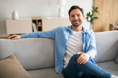 Bir adam iyi aydınlatılmış bir oturma odasında kanepeye oturmuş kameraya bakıyor. Odada bir sehpa, bir kitaplık ve perdeli bir pencere var..