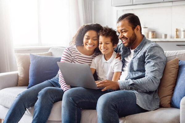 African American family consisting of a father, mother, and two children sitting comfortably on a couch. They are all focused on a laptop screen, engaged in an activity together.