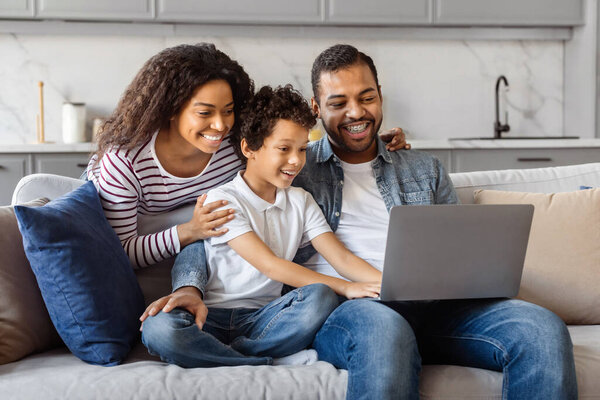 African American family is seated on a couch, all attentively looking at a laptop screen in a living room setting. The laptop is open, displaying a bright screen with various images and text visible