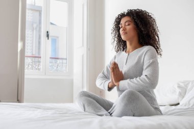 Genç bir İspanyol kadın huzur içinde meditasyon yapıyor. Gözleri kapalı ve elleri dua pozisyonunda. Parlak bir yatak odasında beyaz bir yatakta oturuyor..