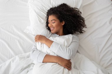 Kıvırcık saçlı İspanyol kadın kabarık bir yastığı kucaklarken mutlu bir şekilde gülümsüyor. Parlak beyaz bir yatak örtüsünün üzerinde rahat bir şekilde yatıyor.