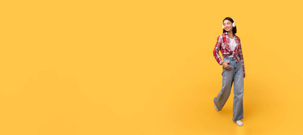 Стильная молодая азиатка в джинсе и клетчатой рубашке уверенно позирует с улыбкой на ярко-желтом фоне