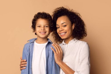 Afro-Amerikalı bir kadın ve bir çocuk birlikte duruyorlar, gülümsüyorlar ve fotoğraf için poz veriyorlar. Kadının kolları çocuğun etrafında, bej rengi arka plan.