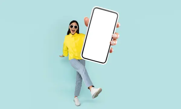 一个戴着太阳镜和一件鲜艳的黄色衬衫的快乐的女人正靠着相机 展示着一个带有空白屏幕的智能手机 准备放置内容 — 图库照片