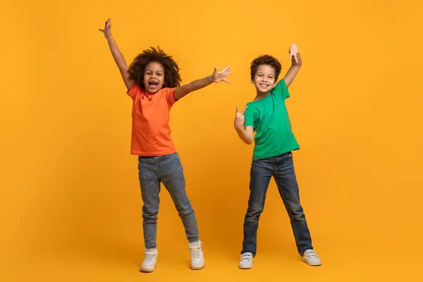 stock image Two young African American siblings are standing with their arms raised in excitement and big smiles on their faces, expressing joy and celebration against a vibrant yellow backdrop.