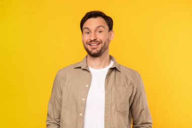 Bir adam parlak sarı bir arka planın önünde dikiliyor. Önüne bakıyor gibi görünüyor, elleri yanda, kameraya gülümsüyor.