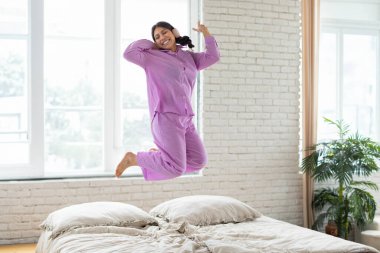 Ortadoğulu bir kadın, parlak bir odada düzgün yapılmış bir yatağın tepesinde enerjik bir şekilde havaya zıplıyor. Kolları açık ve yüzünde neşeli bir ifadeyle orta atlıyor gibi görünüyor..