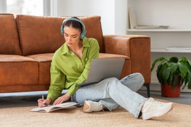 Odaklanmış genç bir kadın sırtı kanepede oturuyor, kulaklık takıyor, dizüstü bilgisayar çalışıyor, not defterine notlar yazıyor, bir çalışma oturumunu veya uzaktan öğrenmeyi işaret ediyor.