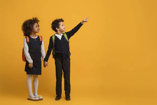 stock image African American young boy and a girl schoolers are pointing at an indiscernible object or direction on yellow background. Their fingers are extended and they are looking intently at the same point.