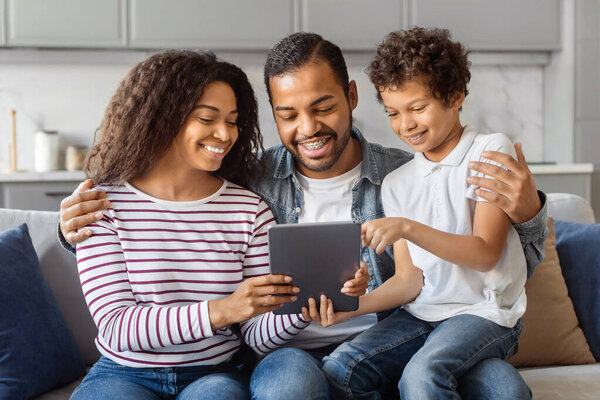 African American man, woman, and child are seated on a couch, engrossed in what appears to be a tablet device. They are focused on the screen, with expressions of concentration and curiosity.