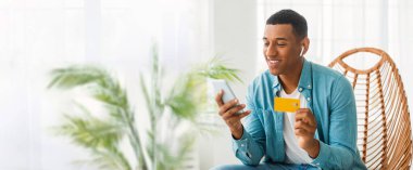 Siyah bir adam heyecan verici bir şekilde akıllı telefonunu ve kredi kartını internet afişi için geniş bir fotokopi alanı olan evde online alışveriş için kullanıyor.