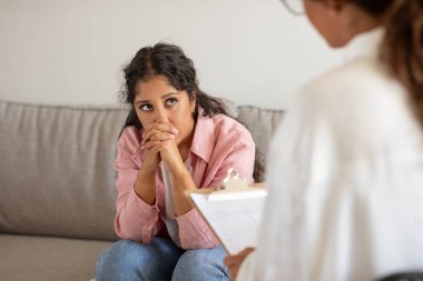 Genç bir kadın bir terapi seansında ellerini birbirine kenetleyerek bir endişe jesti ile koltuğa oturur. Kısmen görülebilen bir terapist elinde bir panoyla karşısında oturuyor.