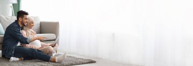 Orta Doğulu hamile çift, birlikte tablet kullanırken rahat bir şekilde dinleniyor ve yan tarafında web pankartı tasarımı için bolca kopya alanı bulunuyor.