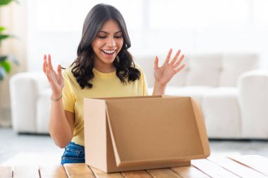 Arap bir kadın açık bir karton kutuya bakarken heyecanlanır, rahat bir ev düzeniyle çevrelenir.