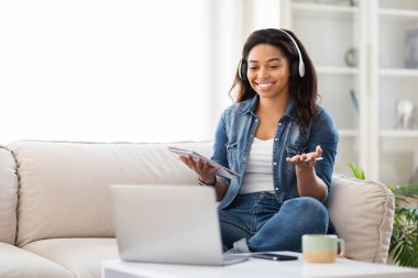 Önünde dizüstü bilgisayar olan bir kanepede oturan Afro-Amerikalı kadın ekrana odaklanıyor ve daktilo ediyor. İşiyle ya da boş zamanlarında meşgul görünüyor..