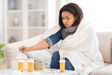 African American woman is sitting on a couch, cocooned in a warm blanket. She looks sick, suffering from cold, take a tissue clipart