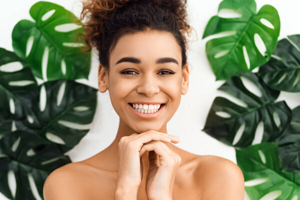 African american woman smiling joyfully with hands held near face in a spa environment, framed by tropical green leaves suggesting freshness