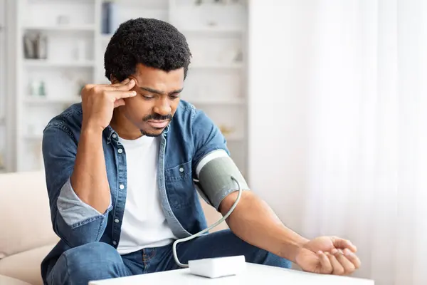 stock image An adult black man looks concerned while using a digital blood pressure monitor at home, suggesting health monitoring or potential issues