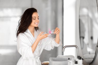 Beyaz bornozlu coşkulu bir milenyum hanımı, iyi donanımlı banyosundaki cilt bakım ürününe hayran.