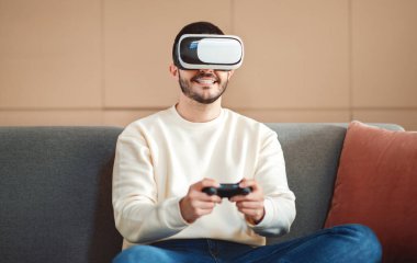 Genç bir adam kafasında sanal gerçeklik kulaklığıyla rahat bir gülümseme ile rahat bir kanepede oturuyor ve sanal gerçeklik oyunundan zevk alıyor gibi görünüyor.