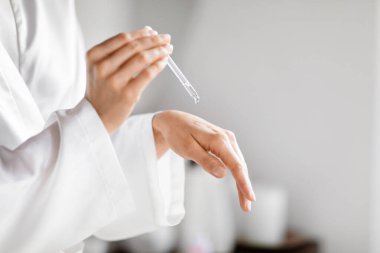 Bornozlu bir kadın banyoda cilt bakım ürünlerini test ediyor, eline serum döküyor.