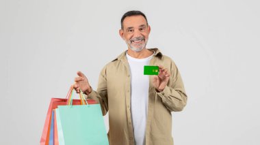 Bir elinde alışveriş poşetleri, diğer elinde kredi kartı olan kıdemli bir adam. Bir alışveriş merkezinde gibi görünüyor, muhtemelen alışveriş yapmaya hazır bir alışveriş merkezi..