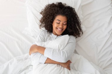 Kıvırcık saçlı neşeli, genç bir İspanyol kadın, güneşli, rahat bir yatakta uzanırken beyaz bir yastığa sarılıyor. Gözleri nazikçe kapalıdır ve ifadesi huzur ve mutluluktur.