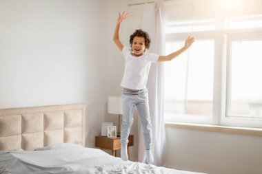 Kıvırcık saçlı genç Afrikalı Amerikalı bir çocuk, iyi aydınlatılmış bir yatak odasında, yatağın üstünde zıplayarak saf neşe ve neşe ifade eden bir yüzle yakalandı.