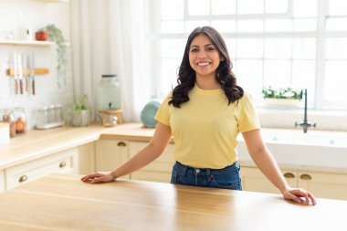 Sarı gömlekli bir ortadoğulu kadın mutfak tezgahında duruyor ve ev işleriyle uğraşıyor. Yemek hazırlarken ya da mutfağı temizlerken odaklanmış ve meşgul görünüyor..